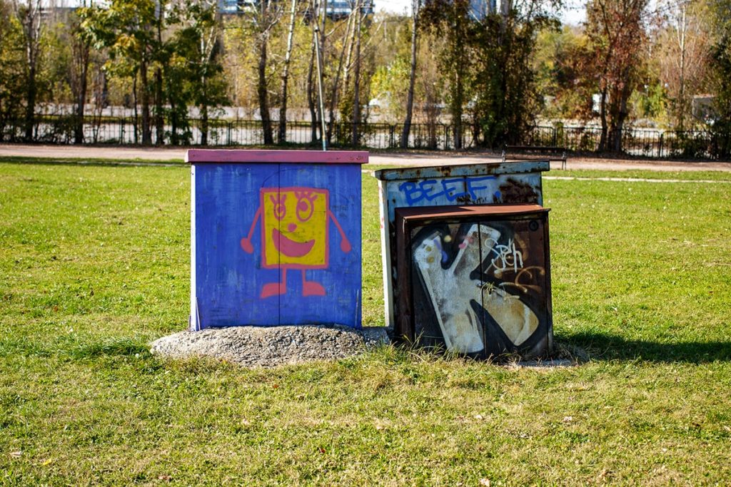 Two electrical boxes covered with graffiti in a sunny park in Sofia, Bulgaria; one features a brightly colored smiling square character, the other has black-and-white stylized letters.