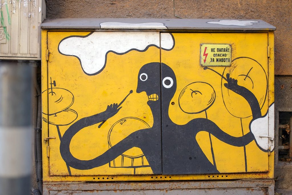 Yellow electrical box with street art depicting a stylized black figure energetically playing drums, accompanied by a warning sign in Cyrillic, in Sofia, Bulgaria.