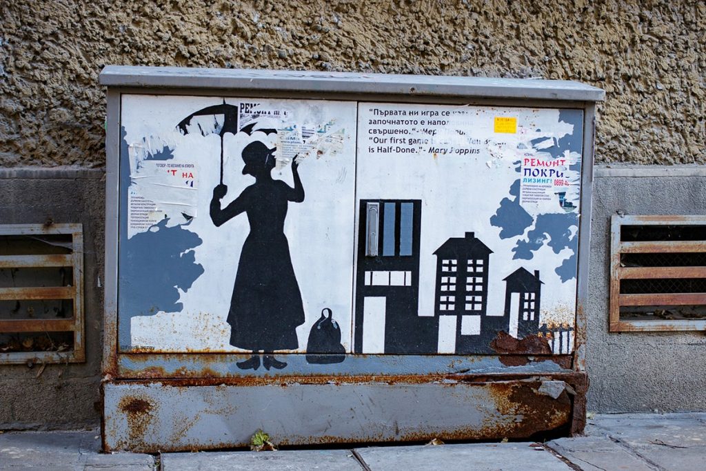 Street art of a Mary Poppins-inspired silhouette holding an umbrella, accompanied by a bilingual quote in Bulgarian and English, painted on an electrical box in Sofia, Bulgaria.