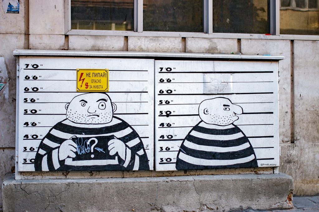 Humorous street art depicting a cartoon prisoner in black-and-white stripes posing for mugshots, painted on an electrical box with a Cyrillic warning sign, in Sofia, Bulgaria.