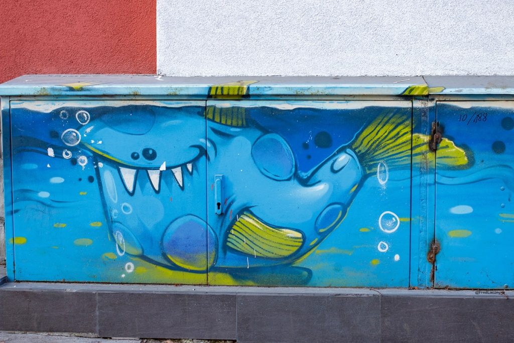 Colorful street art of a playful cartoon-style blue fish with large teeth, swimming and blowing bubbles, painted on an electrical box in Sofia, Bulgaria.