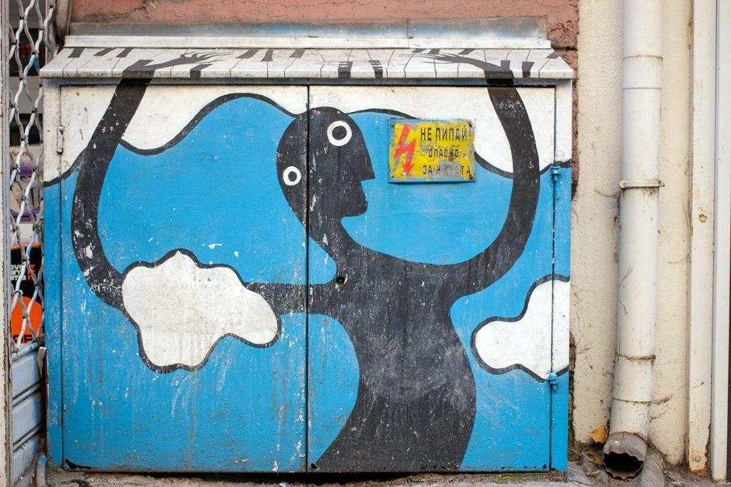 Artistic graffiti showing a black stylized human figure with elongated arms, set against a blue sky background with white clouds, painted on an electrical box in Sofia, Bulgaria.