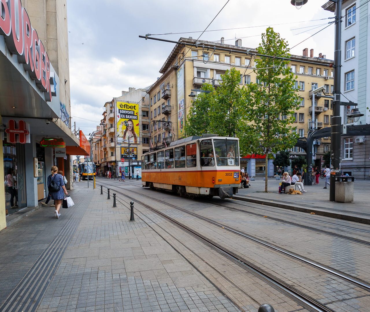 Orange and white tram number 3032 on commercial boulevard with shops and pedestrians in Sofia, Bulgaria, under cloudy sky