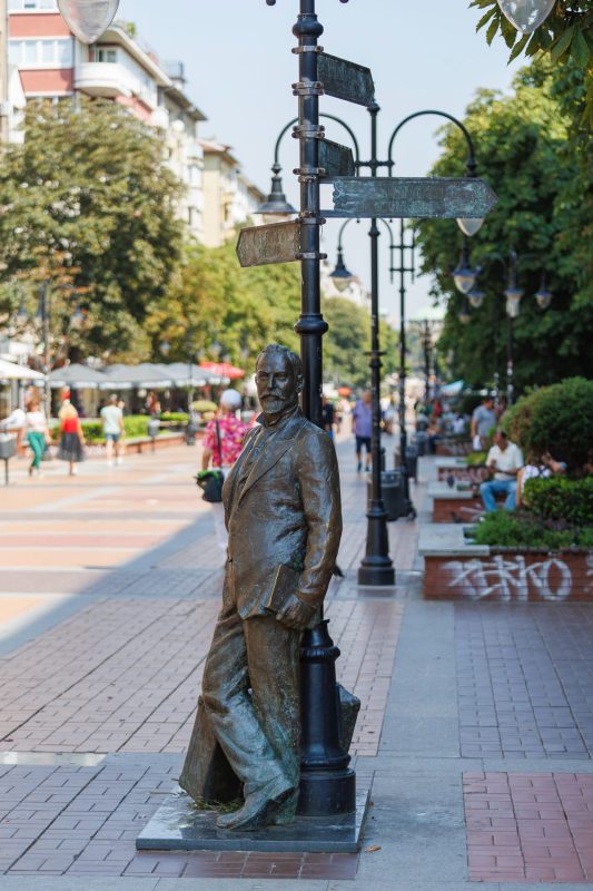 Bronze statue of writer Aleko Konstantinov leaning against a street lamp on Vitosha Boulevard with outdoor cafes and pedestrians in background, Sofia
