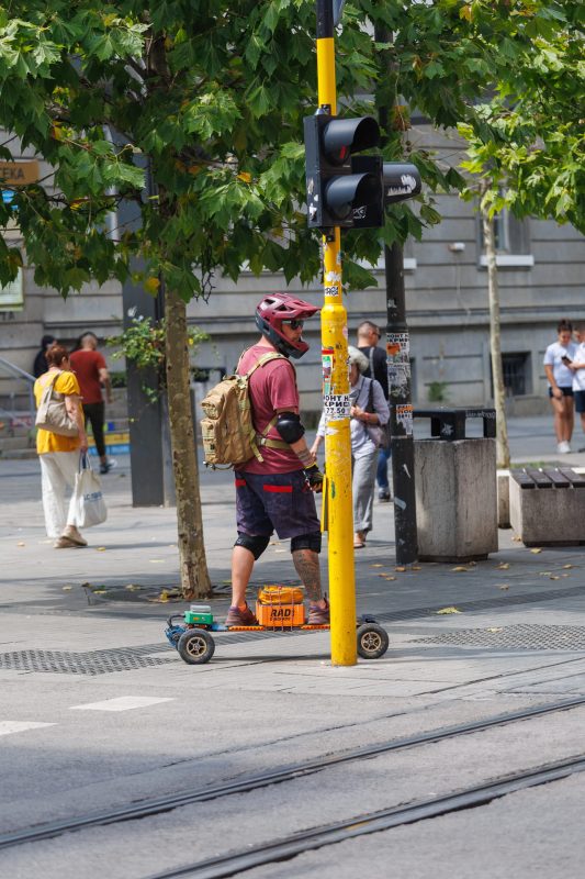 Street performer in cycling gear standing beside yellow traffic light pole with custom RAD cart at pedestrian crossing in Sofia, Bulgaria